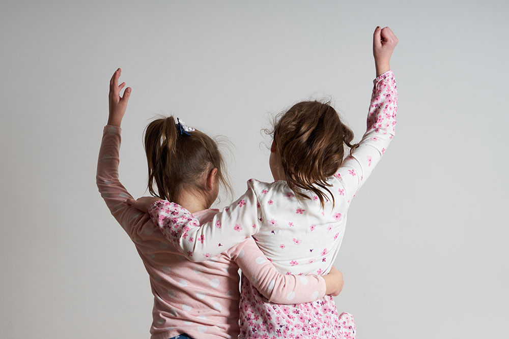 Zwei glückliche Kinder halten sich in den Arm und jeder hebt vor Freude eine Hand in die Luft.